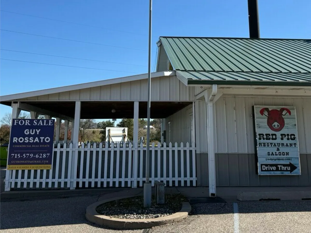 Restaurant building with a green metal roof, white picket fence, Guy Rossato For Sale sign and a roadside billboard at N5696 850th St, Wisconsin